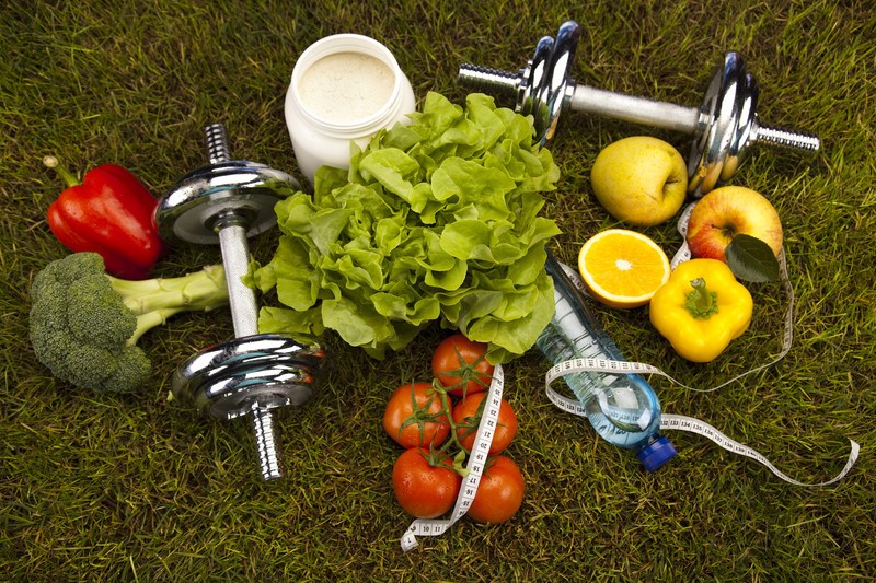 Dumbells, fresh vegetables, a tape measure, and a water bottle rest on a grassy green surface, representing the best foods for weight loss and muscle gain.