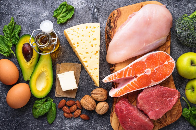 This photo shows an overhead view of several low carb foods, including chicken, salmon, walnuts, cheese, avocado, and eggs, representing the best low carb foods for weight loss.