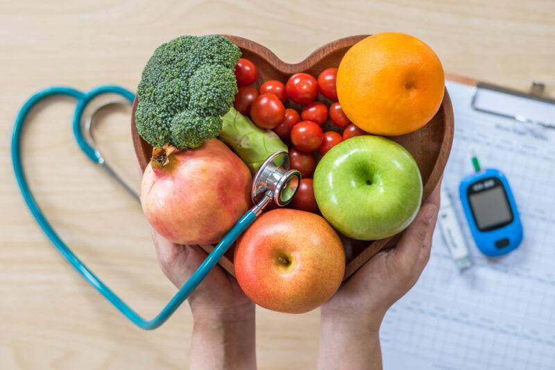 Diabetes,Monitor,,Cholesterol,Diet,And,Healthy,Food,Eating,Nutrition,,World | Food For Net This photo shows an overhead view of two hands holding a heart-shaped bowl of fruits and veggies with a stethoscope above a table with a blood sugar monitor on it, representing the best snack foods for diabetics.