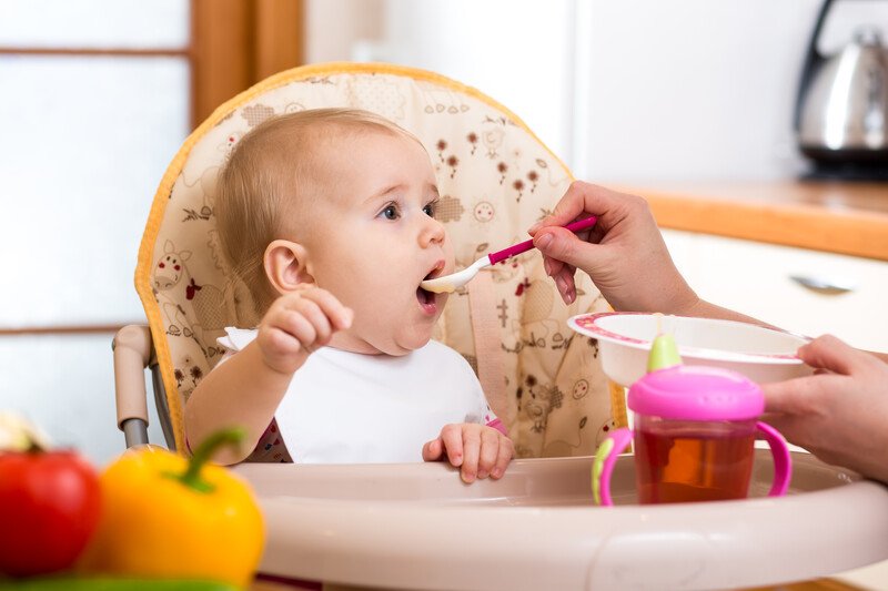 A baby in a high chair takes a bite from a pink and white spoon, representing the best solid foods for babies.