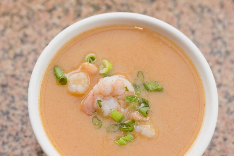 overhead shot of a white bowl full of bisque with herbs and shrimp toppings, resting on a cream colored surface with blue speckles