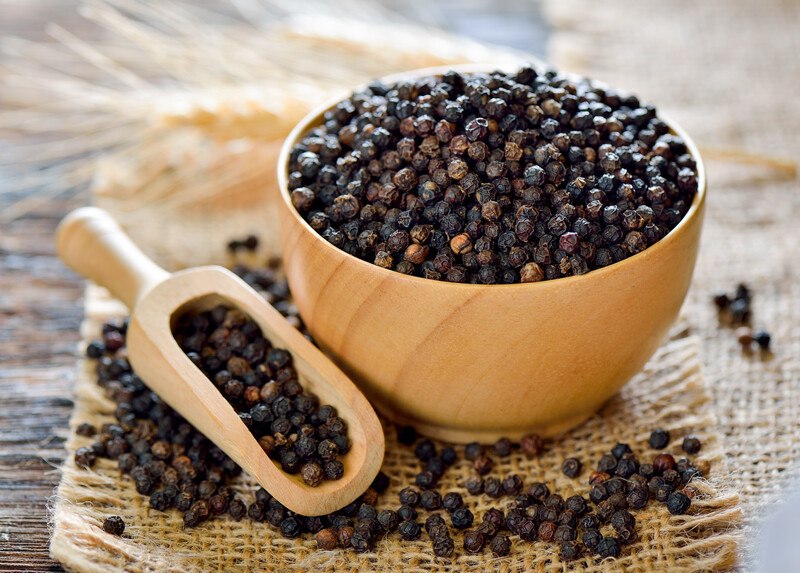 wooden bowl full of black pepper resting on a piece of sack cloth with wooden scoop full of black pepper beside it, and loose black pepper scattered around