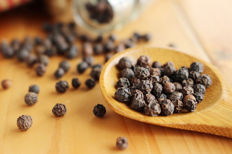 wooden ladle full of black peppers on a wooden surface with lose black peppercorns around it