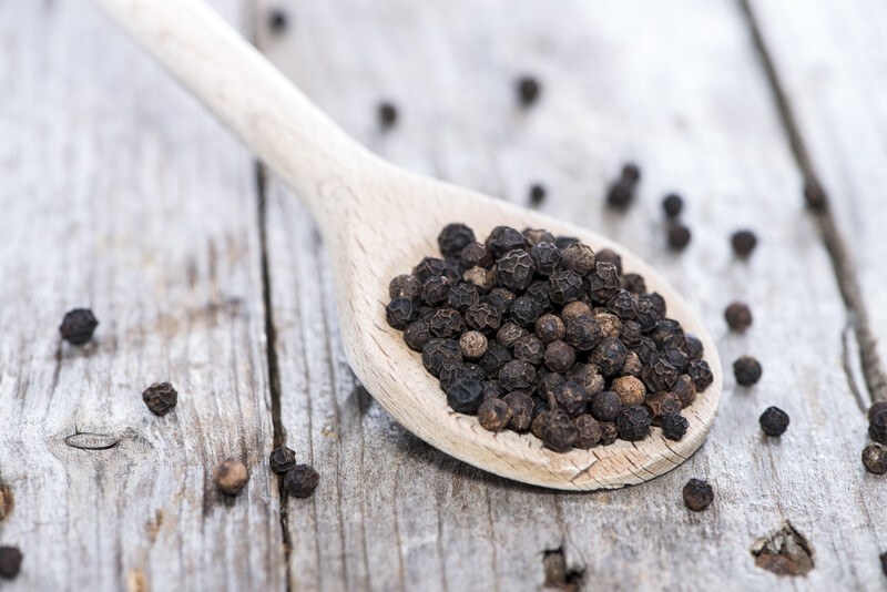 closeup image of a wooden ladle full of black pepper corns with loose black pepper around it on white aged-wooden surface