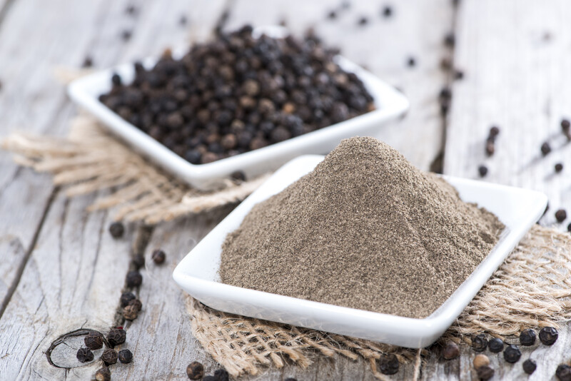 a couple of white dish with whole black pepper and ground black pepper each, on top of a wooden table with scattered whole black peppers on it.