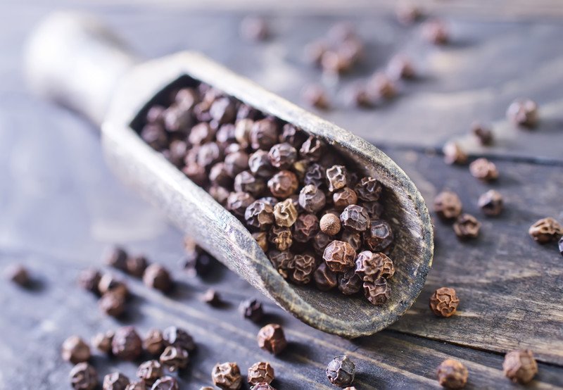 closeup image of a wooden scoop full of black peppercorns on an aged wooden surface with loose black pepper around it