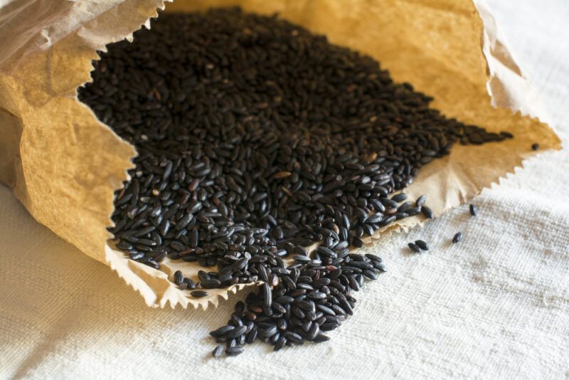 on a tan-colored table cloth is a brown paper bag with raw black rice with a small mound of black rice grains that poured out of the brown paper