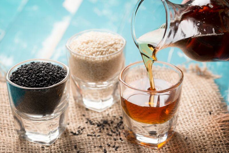 A shot glass of black sesame seeds, another of white sesame seeds, and sesame seed oil being poured into a third shot glass