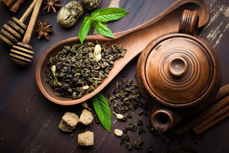 A wooden spoon with black tea leaves, on a table that also has fresh tea leaves, sugar, and a pot of some kind