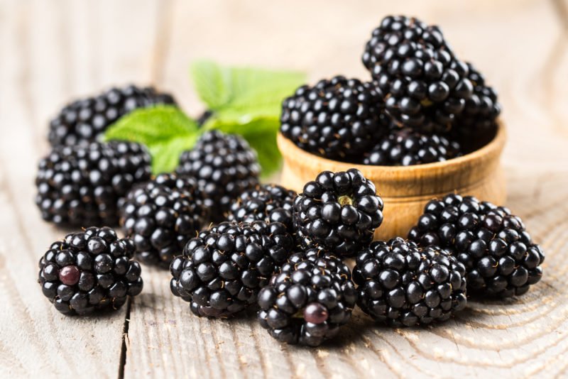 Eine braune Schüssel Brombeeren auf einem Holztisch mit mehr Beeren auf dem Tisch verstreut