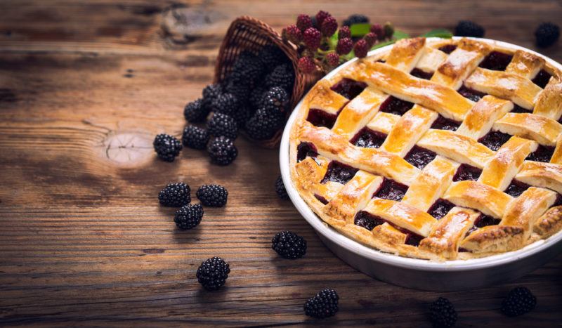 A white dish that contains a blackberry and ginger pie with blackberries scattered on the table next to it