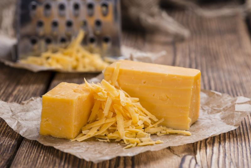 A block of cheddar on paper, some of which has been grated. THere's a grater in the background