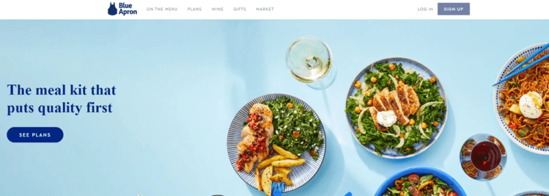 A website screenshot from Blue Apron, showing a light blue table, plus plates and bowls of delicious food and a glass of wine