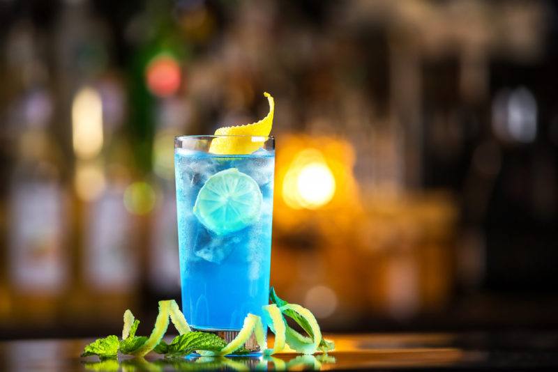 A Blue Lagoon cocktail with ice and a lemon twirl on a bar