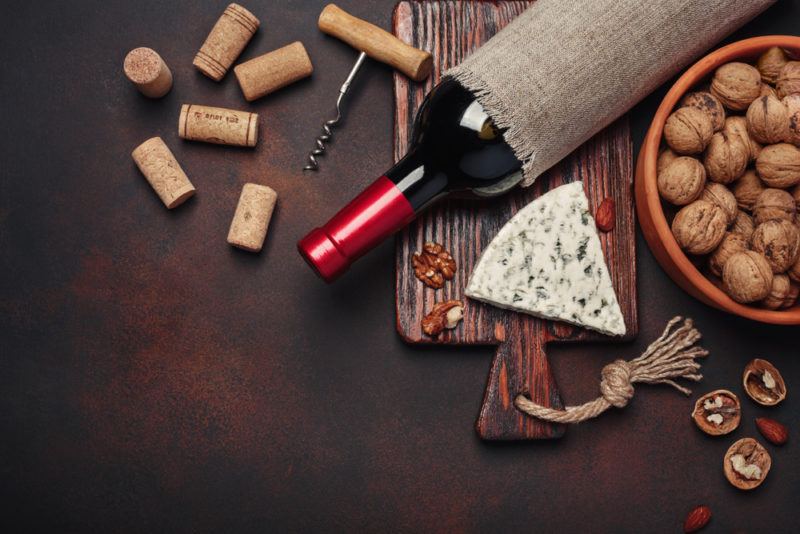 Blue cheese on a wooden board with a few walnuts, next to a bottle of wine, a bowl of whol walnuts, wine corks, a corkscrew and walnut halves