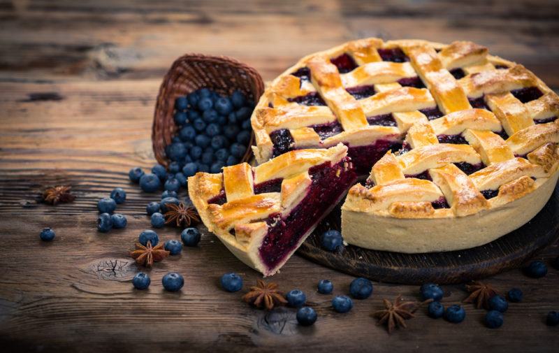 A large blueberry lattice pie with a piece cut out of it, next to a scoop of blueberries