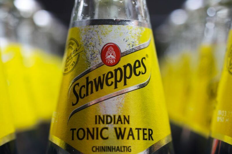 A bottle of Schweppes tonic water in focus, with many other bottles out of focus