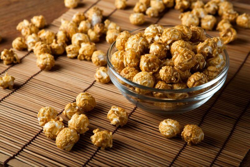 A small glass bowl of caramel popcorn, with plenty more popcorn scattered over the table