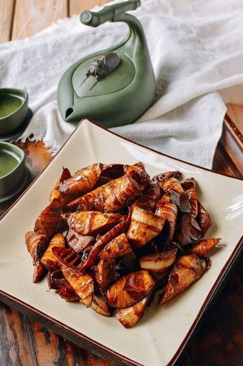 A square white plate containing braised bamboo shoots, next to a green tea pot and some mugs