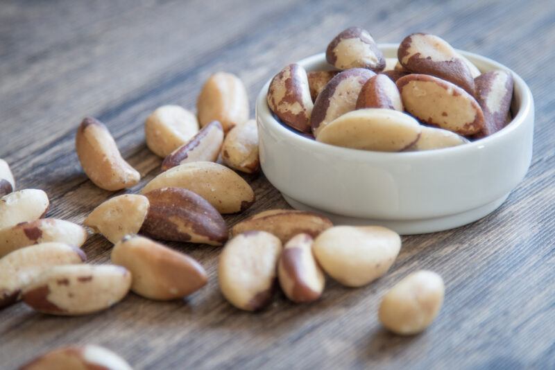 on a wooden surface is a white bowl full of Brazil nuts, with loose Brazil nuts beside it