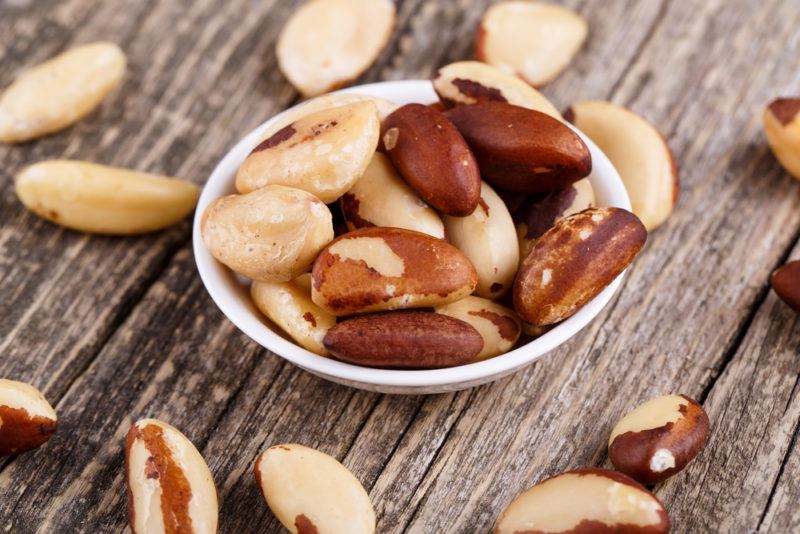 A white bowl with Brazil nuts and other Brazil nuts scattered on a table