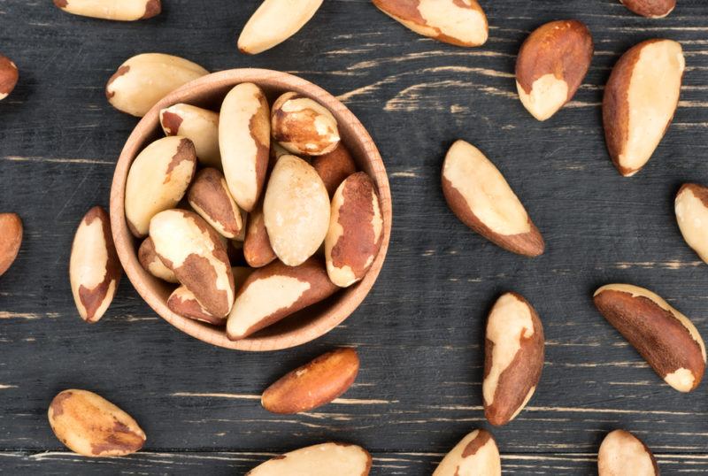 A small wooden bowl with Brazil nuts and more nuts scattered across the table