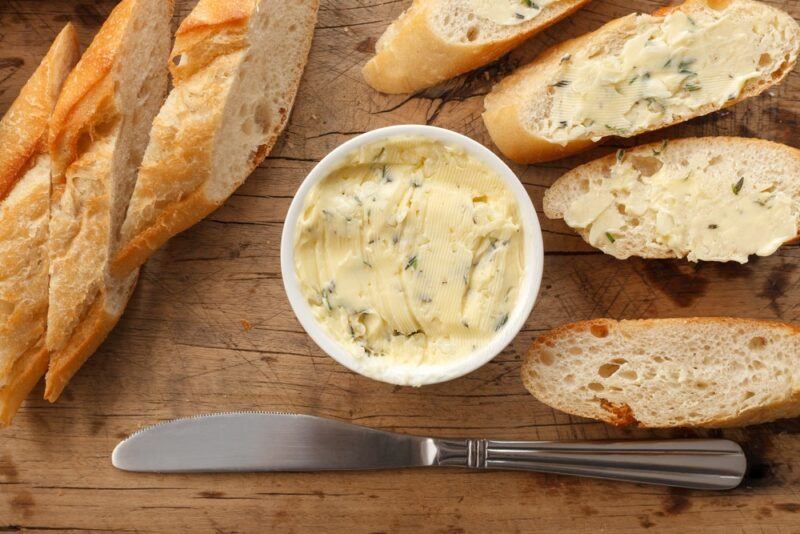 A wooden board with slices of crusty bread, plus a dish of garlic butter. Some of the bread has been spread with butter.