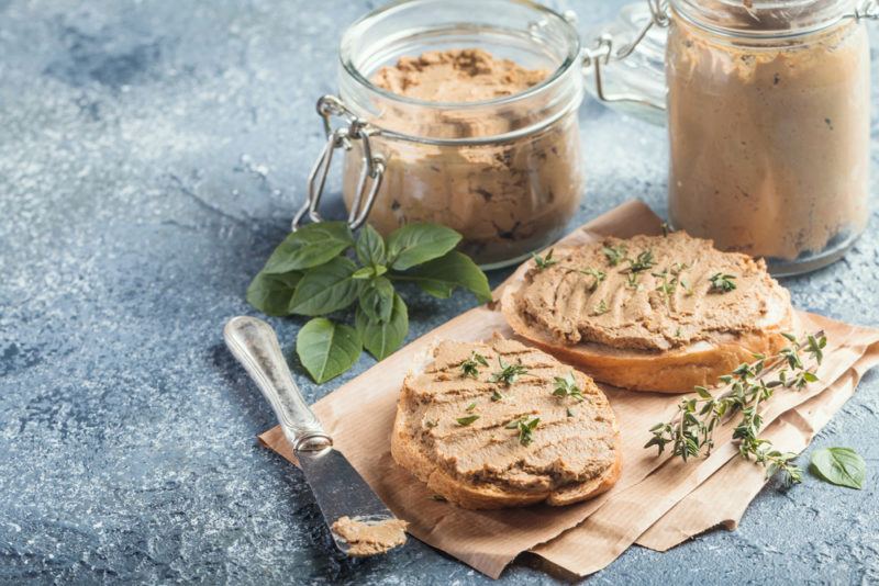 Two pieces of bread on some paper, which have been spread with liver pate. There are two jars of the pate in the background