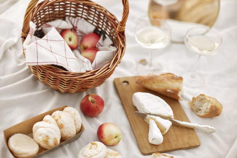 A white picnic mat with bread, cheese, and fruit, along with cutting boards and a basket