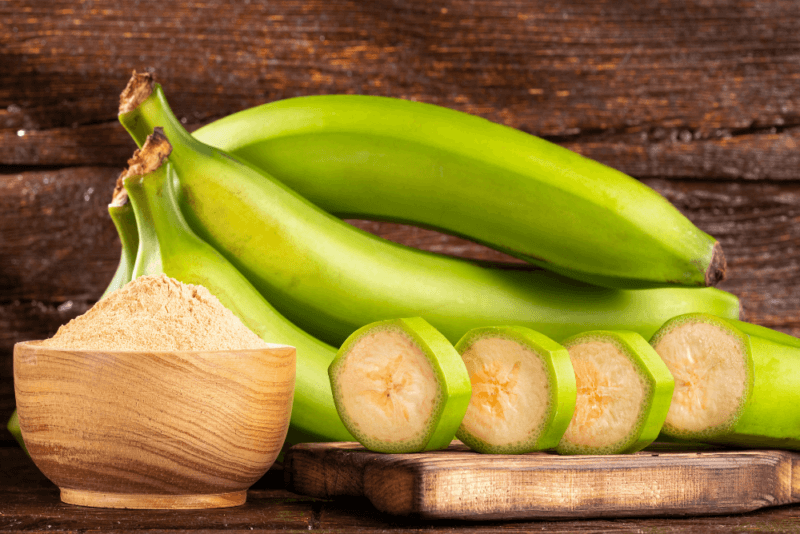 A wooden table with green bananas, some banana slices with their peel still attached, plus a wooden bowl of banana flour