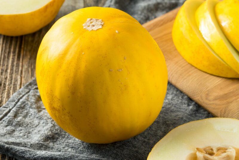 A single large bright yellow canary melon, next to a few slices of the melon on a board