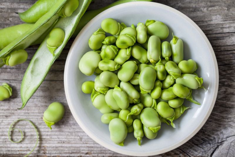 A white bowl of broad beans and some beans in their pods on the table