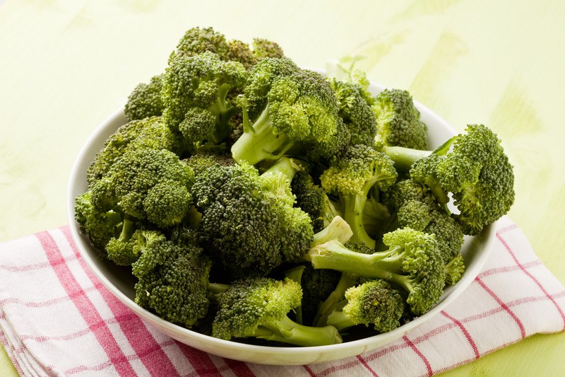 This photo shows a white bowl filled with broccoli florets on a red and white cloth.