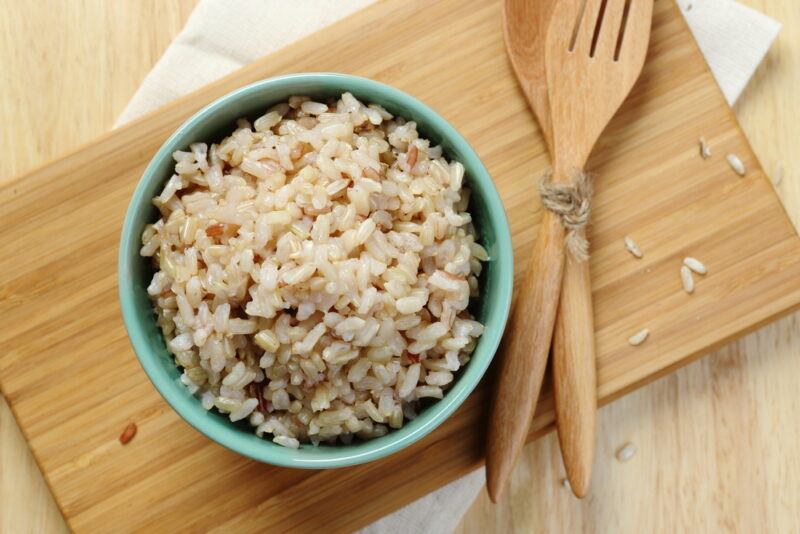 This photo shows an overhead view of a bowl of cooked brown rice with wooden utensils on a bamboo mat, resting on a wooden surface.