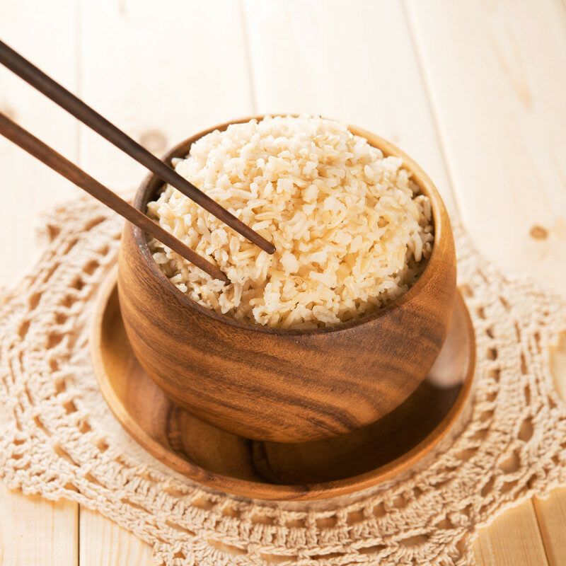 A wooden bowl filled with cooked brown rice and two chopsticks rests on a crocheted doily on a wooden tabletop.