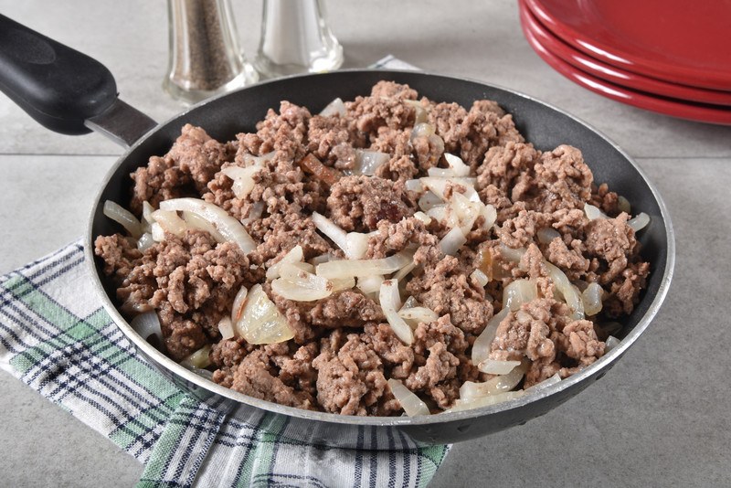 This photo shows a frying pan full of browned ground beef and onions sitting on a kitchen towel on a white countertop.