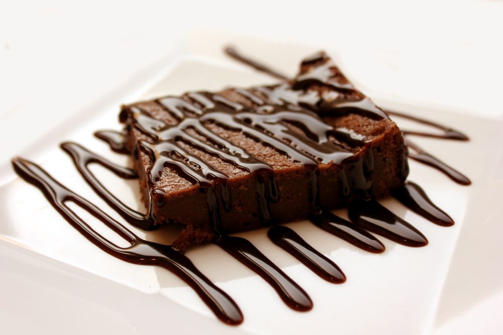 Chocolate brownie with chocolate sauce drizzled diagonally across the brownie and square white plate it sits on 