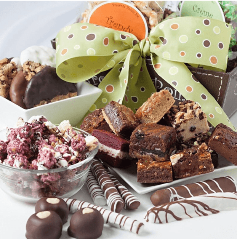 An array of sweet treats including a pile of brownie bites on a white plate, bon bons, chocolate covered pretzels, cookies in a white rectangle bowl, a glass bowl of flavored gourmet popcorn, and a bow with goodies in the background featuring a green polka dotted bow