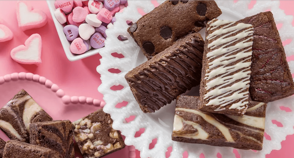 A variety of Valentine's day brownie bars and squares surrounded by valentines candy on a pink backdrop. 5 brownie bars are on a white tray flavors include chocolate, chocolate chip, and chocolate with white chocolate drizzle.  Left lower corner on a pink heart shaped plate are brownie squares