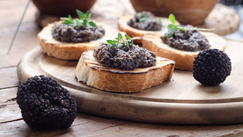 A wooden board with four slices of bruschetta, each topped with black truffle paste