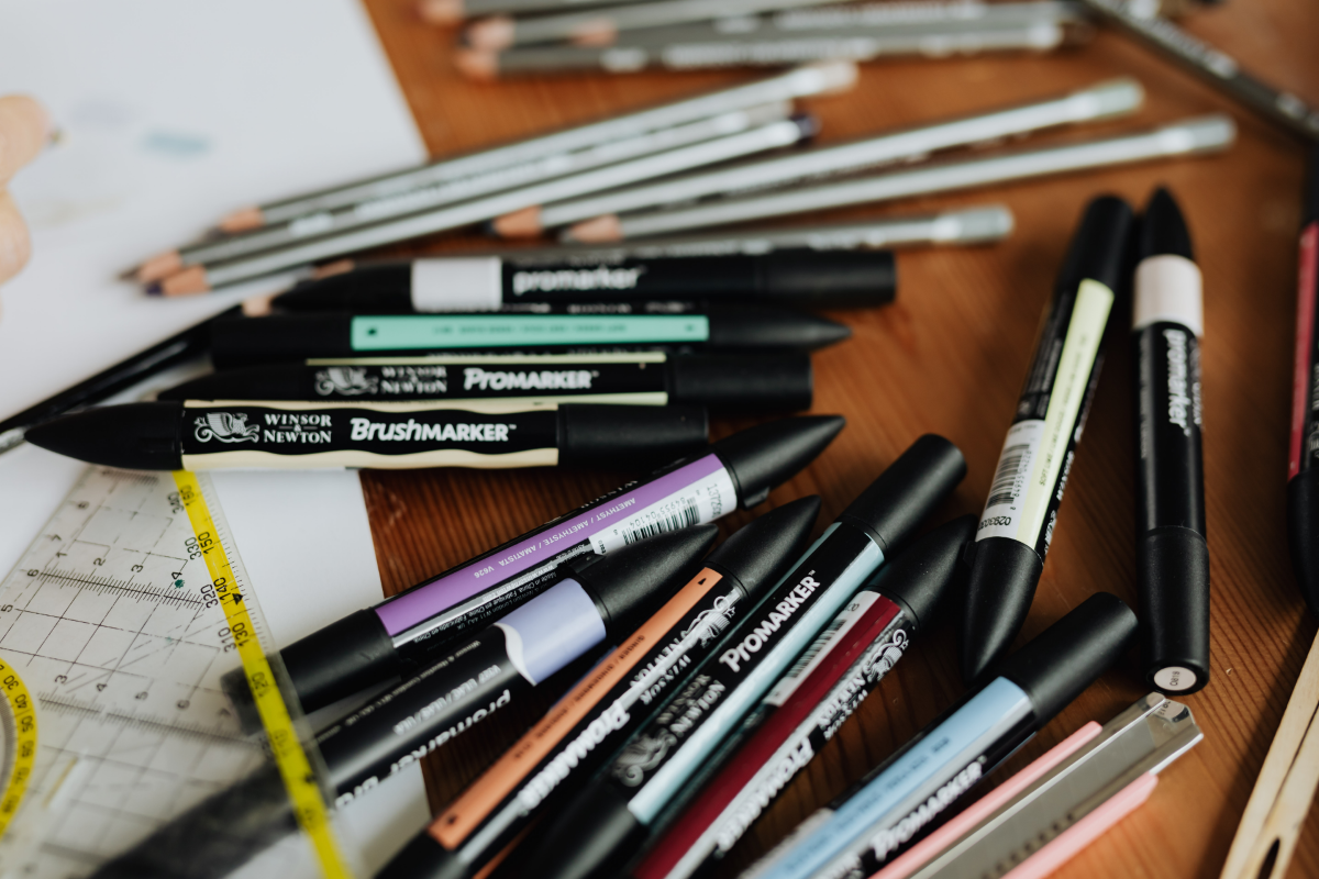 Brush markers, rulers, and colored penciles along with other art supplies scattered on a wood table.