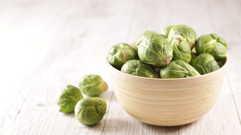 on a white wooden surface is a wooden bowl full of Brussels sprouts, with 3 loose Brussels sprouts beside it