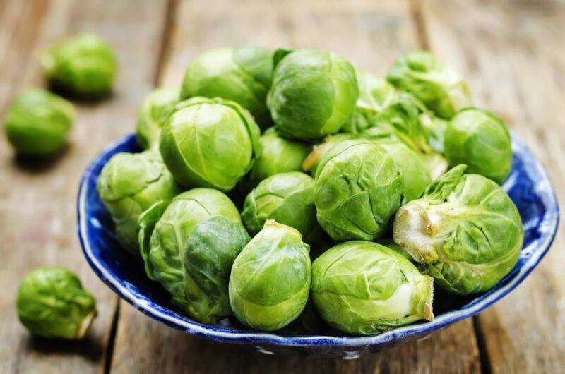 on a rustic looking wooden surface is a blue ceramic bowl full of Brussels sprouts, with loose Brussels sprouts beside it