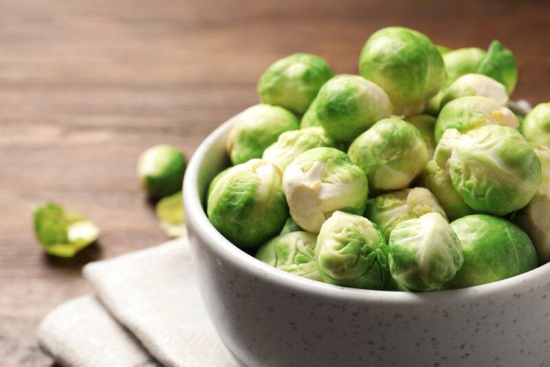 on a wooden surface is a white bowl full of Brussels sprouts resting on a white table napkin