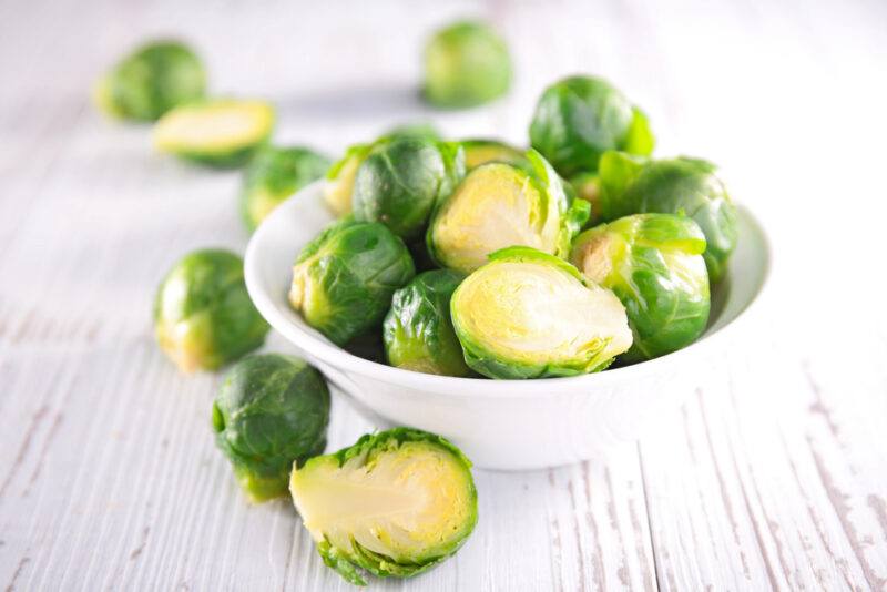 on a white wooden surface is a white bowl full of Brussels sprouts, around it are loose Brussels sprouts