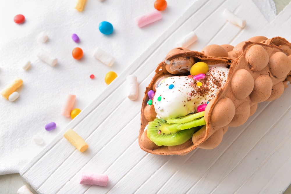 A white table with a bubble waffle that has been filled with sweet ingredients, with various toppings scattered on the table