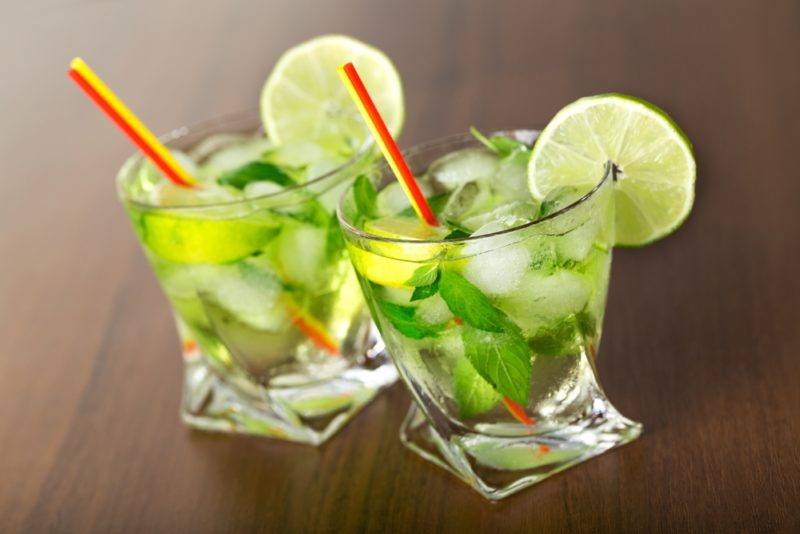 Two twisted glasses containing a Caipiroska cocktail with ice, limes, straws and leaves