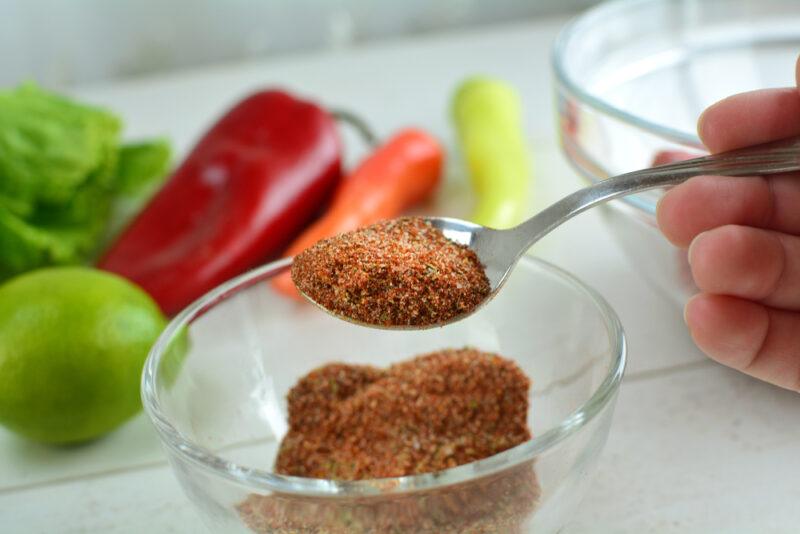 partial image of a hand holding a spoon full of Cajun seasoning over a clear glass bowl of Cajun seasoning, behind are fresh veggies like bell peppers, carrots, greens, and peppers