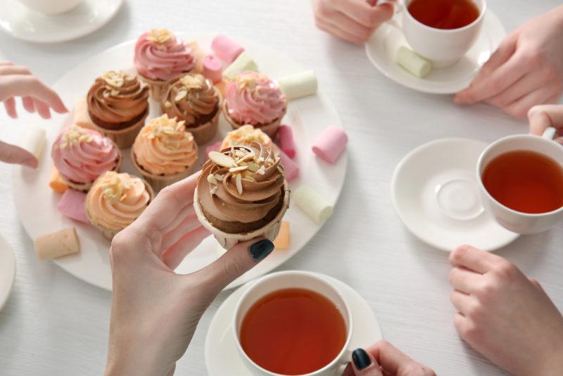 A white plate of cakes with people sitting around it holding mugs of tea. One hand is lifting one of the cakes