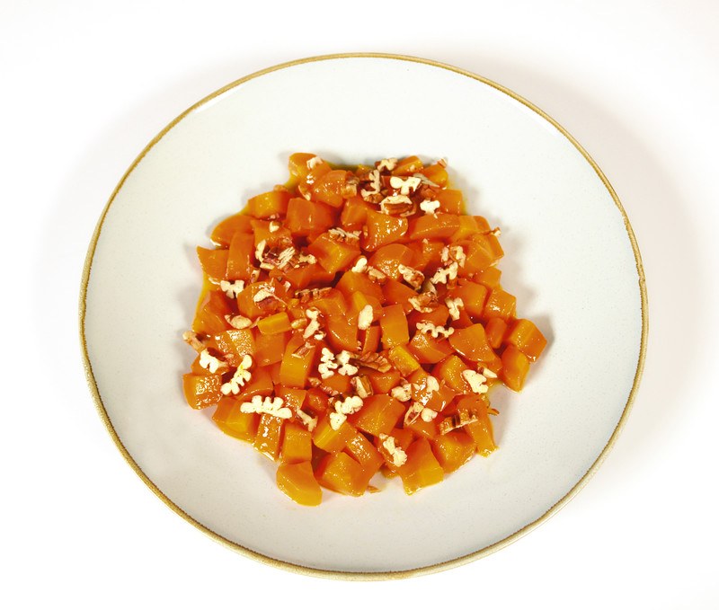 This photo shows an overhead view of a white plate of cooked diced carrots against a white background.